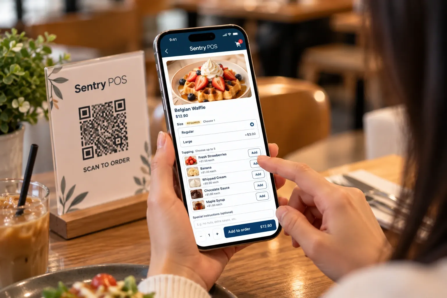 A customer at a café table using their phone to choose options for a Belgian waffle on the Sentry POS self-ordering menu, beside a ‘Scan to order’ QR code sign.